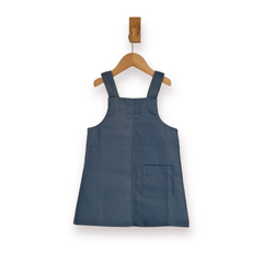 A slate blue sleeveless pinafore-style dress with shoulder straps and a front patch pocket, hanging on a wooden hanger against a plain white background.