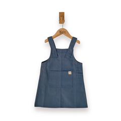 A slate blue pinafore dress with shoulder straps and a front patch pocket, hanging on a wooden hanger ].