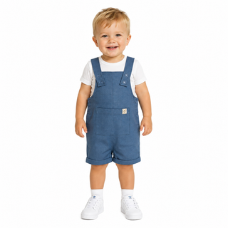 A smiling toddler standing against a white background, wearing blue dungaree shorts over a white t-shirt and white trainers