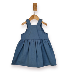 A blue girls dungaree dress with shoulder straps and a gathered skirt, hanging on a wooden hanger from a wall hook against a white background.