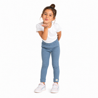A young girl with her hair in a bun stands against a white background, wearing a white t-shirt, light blue slim fit leggings, and white trainers, blowing a kiss.