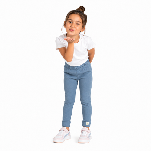 A young girl with her hair in a bun stands against a white background, wearing a white t-shirt, light blue slim fit leggings, and white trainers, blowing a kiss.