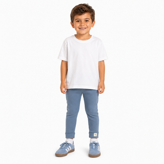 A young boy standing against a plain white background, wearing a white short-sleeve t-shirt, blue pants, and blue trainers. 