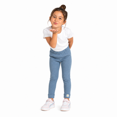 A young girl with her hair in a bun stands against a white background, wearing a white t-shirt, light blue slim fit leggings, and white trainers, blowing a kiss.