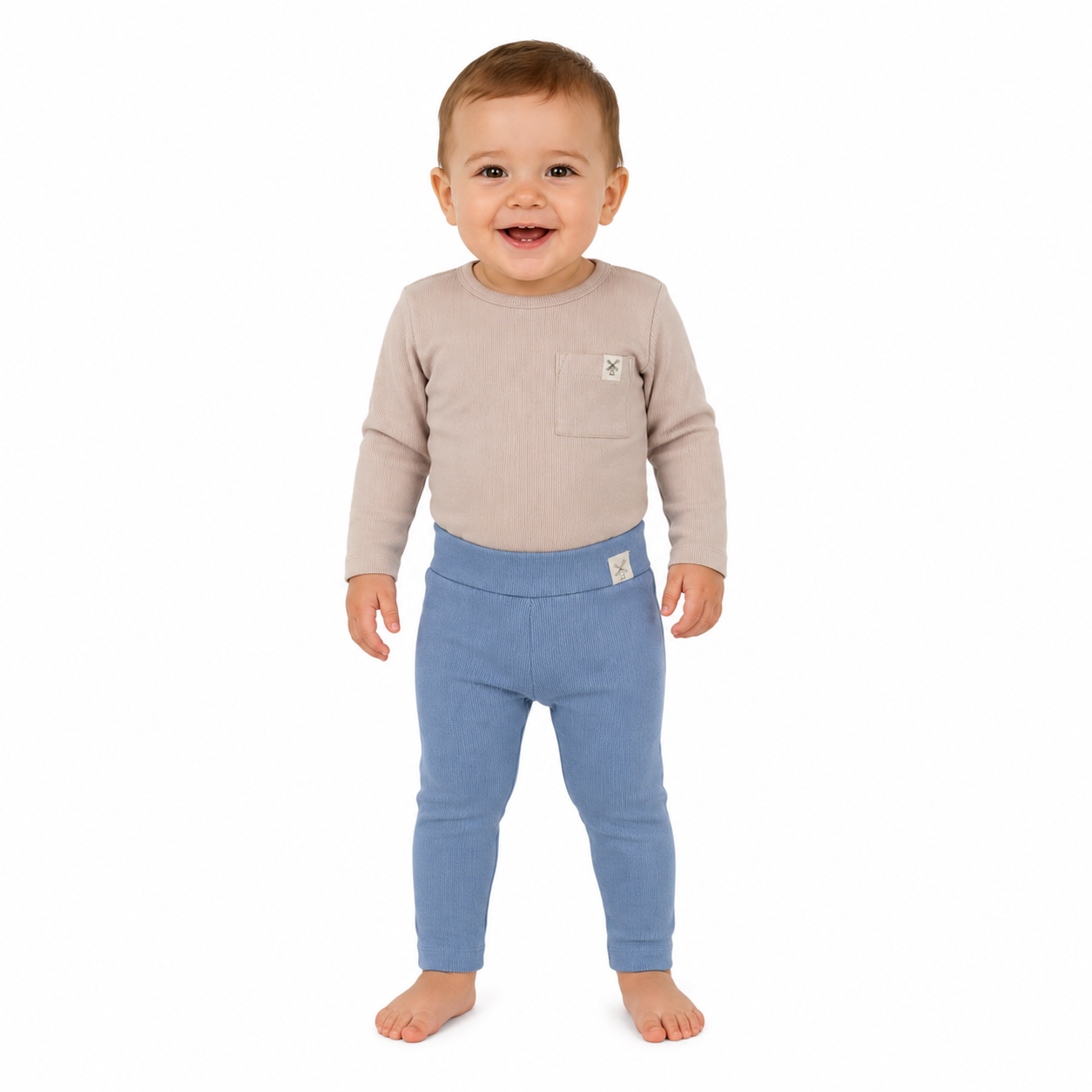 A smiling toddler standing barefoot against a plain white background, wearing a light beige long-sleeve top with a chest pocket and blue ribbed leggings.
