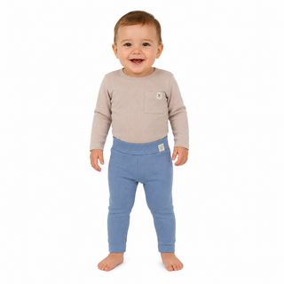 A smiling toddler standing barefoot against a plain white background, wearing a light beige long-sleeve top with a chest pocket and blue ribbed leggings.