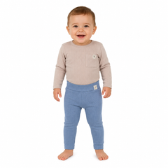 A smiling toddler standing barefoot against a plain white background, wearing a light beige long-sleeve top with a chest pocket and blue ribbed leggings.
