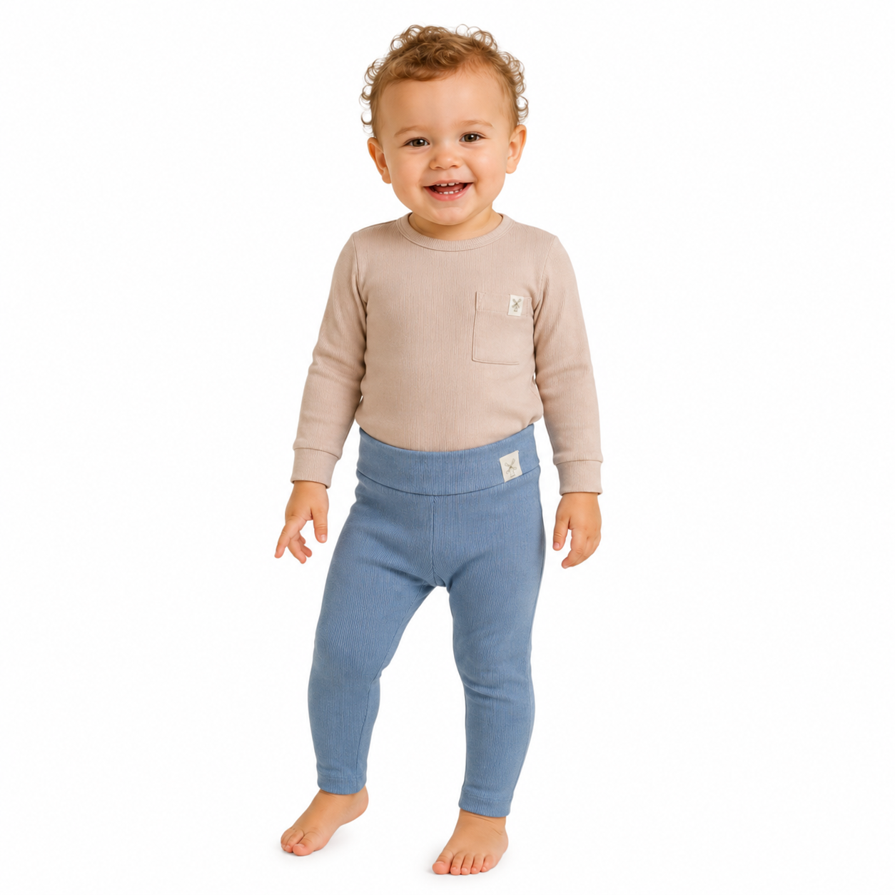 A smiling girl standing barefoot against a plain white background, wearing a light beige long-sleeve top with a chest pocket and blue leggings.