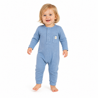 A toddler with blonde hair wearing a light blue long-sleeve footless one-piece romper with a three-button placket and a small chest pocket, standing against a plain white background.