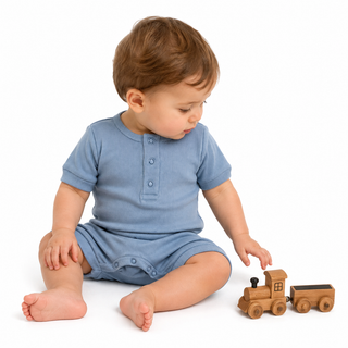 A small boy sitting on a white studio background wearing a blue short-sleeve romper, looking down at a small wooden toy train.