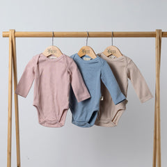 Long-sleeved body suits in pink, blue and oat, hanging from wooden frame