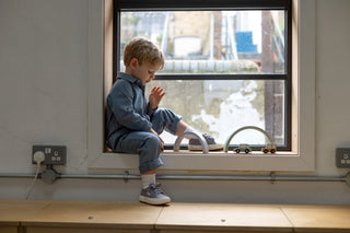 Boy sat in window wearing blue boilersuit playing with wooden toy cars