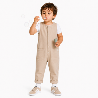 A toddler boy wearing a beige corduroy overall jumpsuit over a white t-shirt, standing and blowing bubbles against a clean white background.