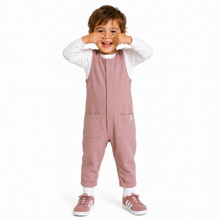 A toddler standing against a white background, wearing dusty rose overall with a white long-sleeve shirt underneath and matching Adidas trainers.