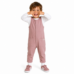 A toddler standing against a white background, wearing dusty rose overall with a white long-sleeve shirt underneath and matching Adidas trainers.