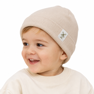 A smiling toddler wearing a ribbed beige knit beanie with a small white square patch featuring a green windmill logo, photographed against a clean white background.