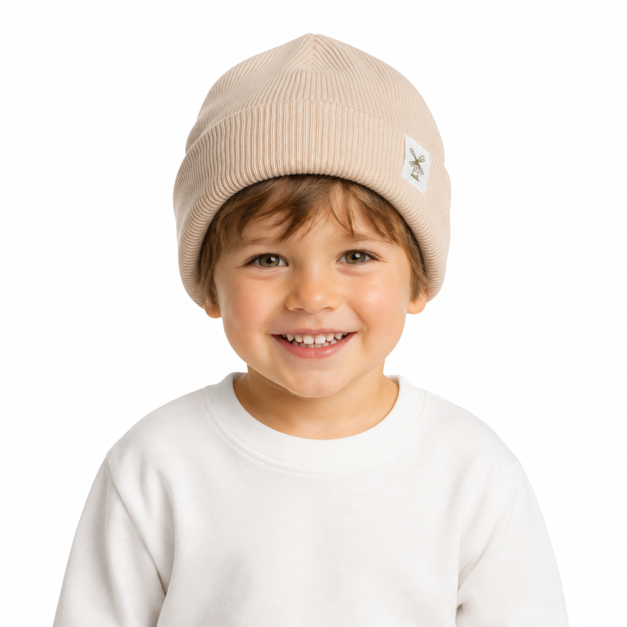 A smiling young child wearing a light beige ribbed knit beanie with a small embroidered logo patch on the side, paired with a white crewneck sweatshirt against a plain white background.