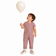 A toddler stands against a plain white background holding a cream-colored balloon. The child is wearing a pink short-sleeve one-piece jumpsuit with front pockets and sneakers.