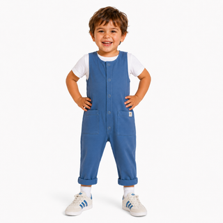 A smiling toddler stands with hands on hips wearing blue overall-style pants over a white short-sleeve shirt, with white sneakers, on a light gray studio background.