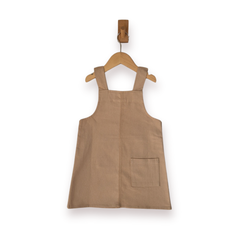 A sleeveless ecru pinafore-style dress with shoulder straps and a front patch pocket, hanging on a wooden hanger against a plain white background.