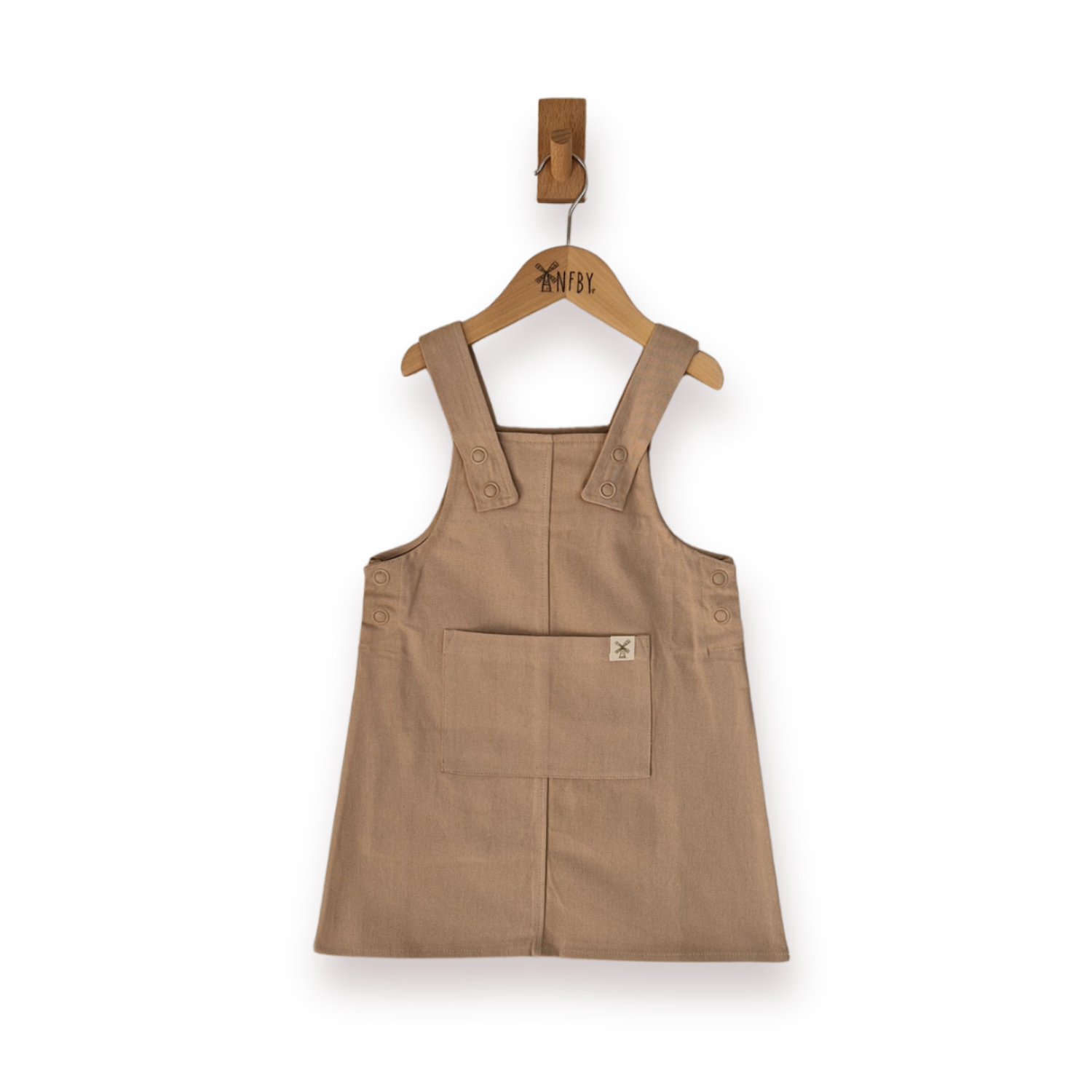 A sleeveless ecru utility pinafore style dress with a front patch pocket, hanging on a wooden hanger, set against a plain white background.