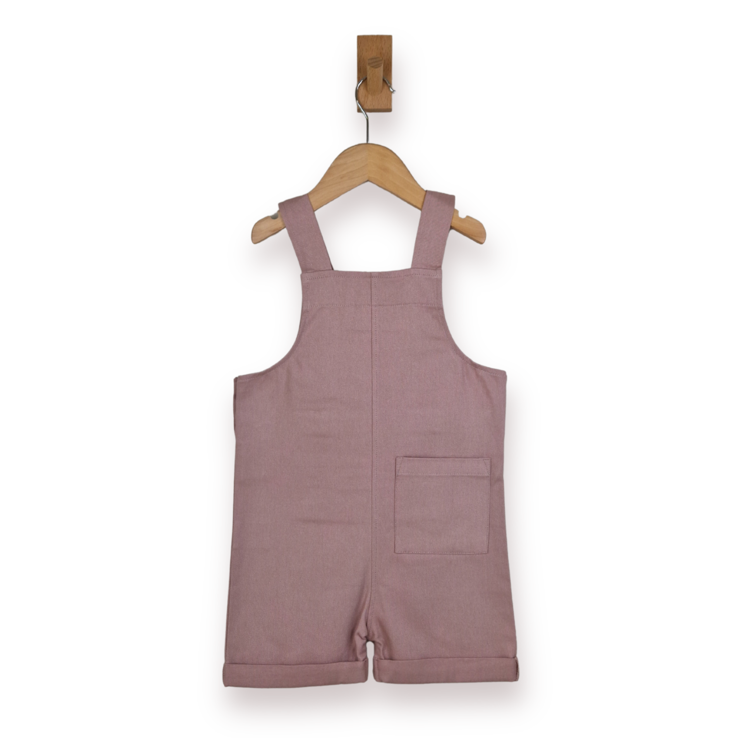 A dusty rose pink cotton short dungarees for toddlers, hanging on a wooden hanger from a wall hook against a white background.