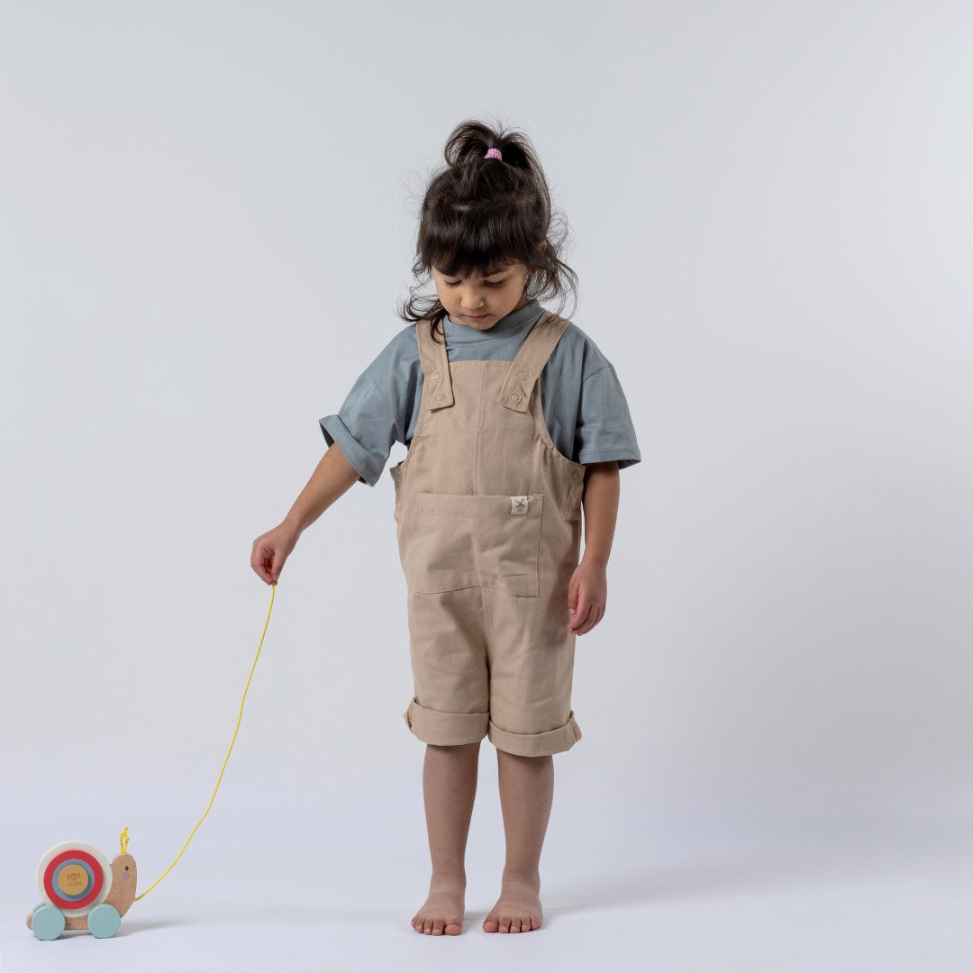 Beige dungaree shorts with shoulder straps and front pocket, being modelled by young girl with wooden toy snail