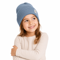 A young girl with light brown hair wearing a dusty blue ribbed knit beanie and a cream-colored long-sleeve top, posing against a solid white background.