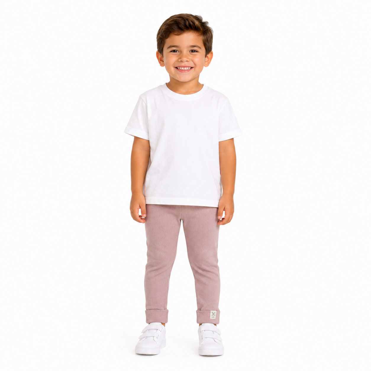 A young boy smiling and standing against a plain white background, wearing a white short-sleeve t-shirt, dusty pink pants, and white trainers.