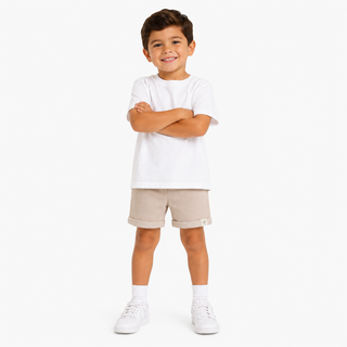 A young boy modeling a white short-sleeve crewneck t-shirt and beige rolled-calf shorts, paired with white socks and trainers, standing against a white studio background.
