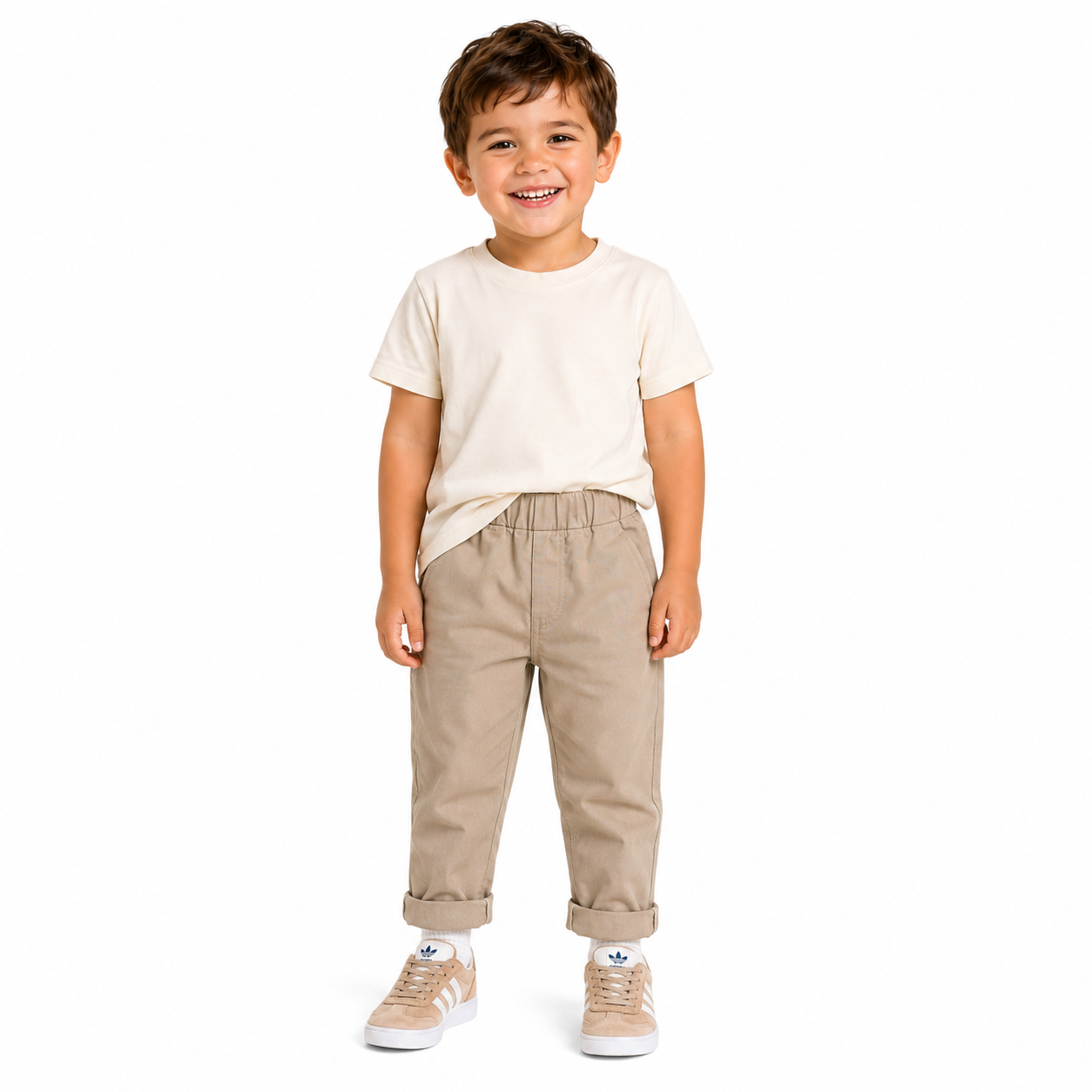 A young boy smiling and standing against a white background, wearing a cream short-sleeve t-shirt, beige elastic-waist pants, and beige sneakers.