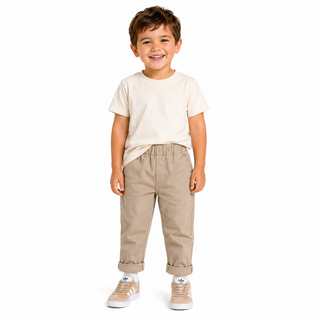 A young boy smiling and standing against a white background, wearing a cream short-sleeve t-shirt, beige elastic-waist pants, and beige sneakers.