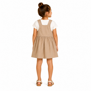 A young girl with her hair in a bun is shown from the back, wearing a beige dungaree dress over a white short-sleeve t-shirt and beige sandals, against a plain white background.