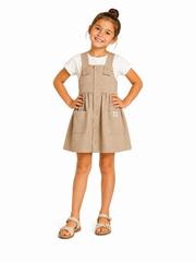 A young girl with her hair in a bun stands with hands on hips wearing a beige linen-look pinafore dress over a white t-shirt and tan sandals, photographed against a solid white background.