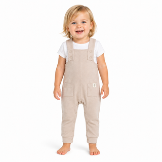 A smiling toddler standing barefoot against a plain white studio background, wearing light beige overall-style pants over a white short-sleeve top.