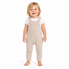 A smiling toddler standing barefoot against a plain white studio background, wearing light beige overall-style pants over a white short-sleeve top.