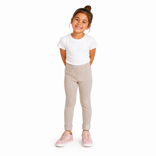 A young girl with her hair in a bun stands against a plain white background, wearing a white short-sleeve t-shirt, light beige jogger pants, and pink sneakers.