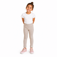 A young girl with her hair in a bun stands against a plain white background, wearing a white short-sleeve t-shirt, light beige jogger pants, and pink sneakers.