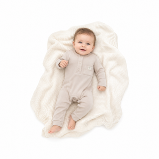 A baby lying on a textured cream blanket, wearing an ecru long-sleeve romper with a button placket and a small chest patch.