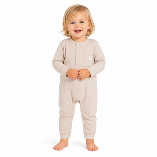 A smiling toddler with blonde hair stands barefoot against a white background, wearing a light beige long-sleeve footless one-piece romper with a small white embroidered logo on the left chest.