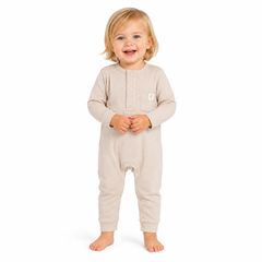 A smiling toddler with blonde hair stands barefoot against a white background, wearing a light beige long-sleeve footless one-piece romper with a small white embroidered logo on the left chest.