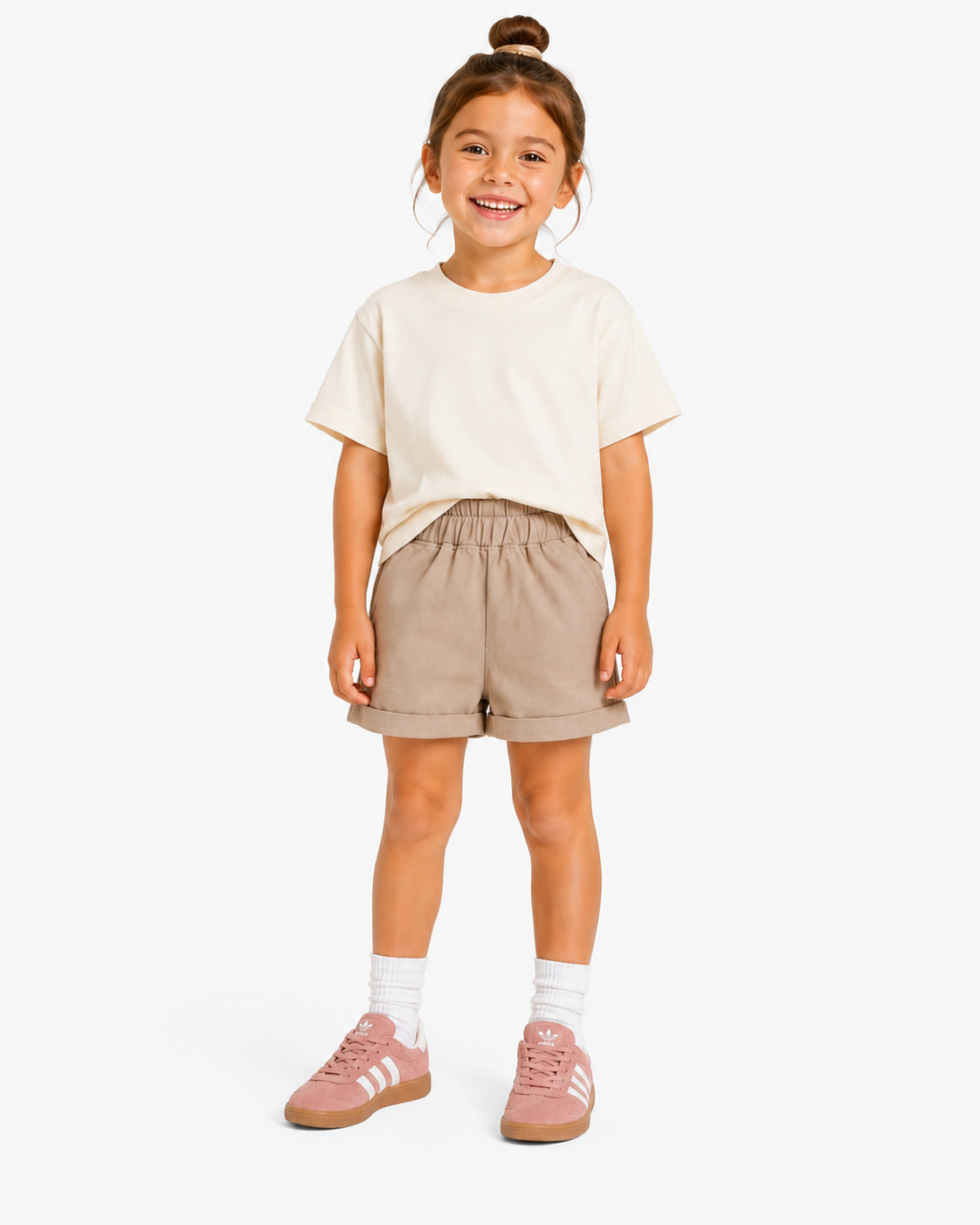 A young girl stands against a light studio background wearing a cream short-sleeve t-shirt, beige shorts, white socks, and pink sneakers.