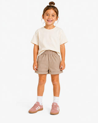 A young girl stands against a light studio background wearing a cream short-sleeve t-shirt, beige shorts, white socks, and pink sneakers.