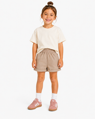 A young girl stands against a light studio background wearing a cream short-sleeve t-shirt, beige shorts, white socks, and pink sneakers.