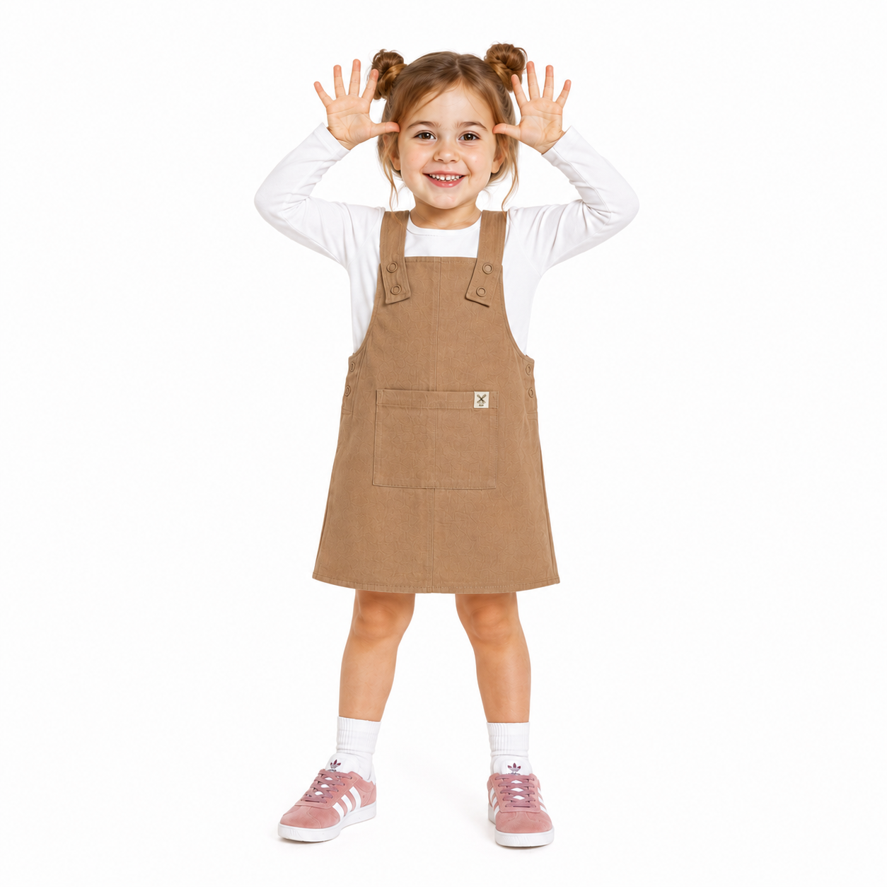 A young girl with brown pigtails stands against a white background wearing an oat dungaree dress, over a white long-sleeve shirt, paired with pink sneakers and white socks.