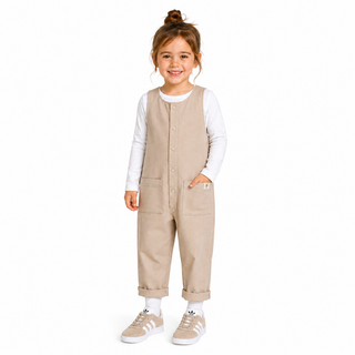 A young girl stands against a plain white background wearing a beige corduroy jumpsuit over a white long-sleeve shirt, with beige sneakers and white socks.