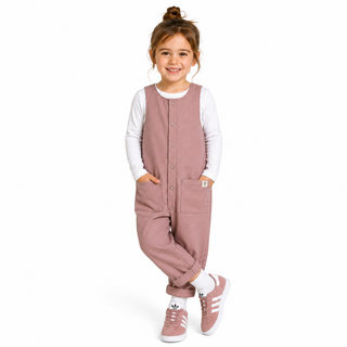 A young girl with her hair in a bun stands against a white studio background wearing a dusty rose corduroy overall jumpsuit over a white long-sleeve shirt, paired with white socks and matching pink trainers.