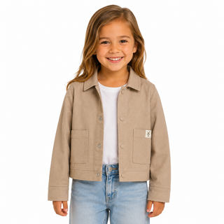 A young girl smiling and wearing a beige shacket over a white t-shirt and blue jeans, photographed against a solid white background.