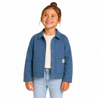 A young girl with her hair in a bun smiling and looking up, wearing a blue button-up jacket over a cream t-shirt and light blue jeans, photographed against a solid white background.