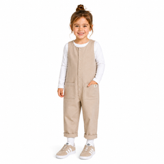 A young girl stands against a plain white background wearing a beige corduroy jumpsuit over a white long-sleeve shirt, with beige sneakers and white socks.